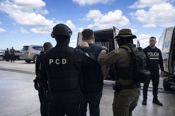 Un hombre es escoltado por dos agentes con equipo táctico. Alrededor hay más personal policial y vehículos bajo un cielo azul con nubes blancas.