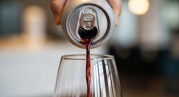Mano humana vertiendo vino tinto de lata plateada con abrelatas en copa de cristal transparente.
