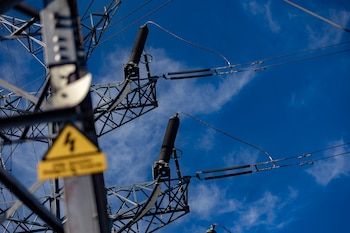 Torre y cables de alta tensión junto a una señal de peligro eléctrico bajo un cielo azul.