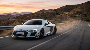 Audi R8 blanco deportivo conduciendo en una carretera de montaña al atardecer. Se ve polvo detrás del coche y montañas en el fondo.