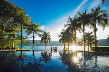 Piscina infinita reflejando cielo azul y palmeras, con tres personas sentadas y una persona saltando, frente a una laguna y montañas con sol