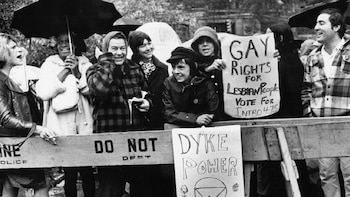 Fotografía en blanco y negro de Marsha P. Johnson y Sylvia Rivera sonriendo bajo paraguas, junto a otros manifestantes LGBTQ+ que sostienen pancartas en una protesta