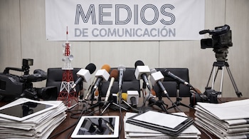 Mesa de prensa con micrófonos, tabletas, periódicos, cámaras de video, una cámara de fotos, una torre de radio en miniatura y una pancarta.