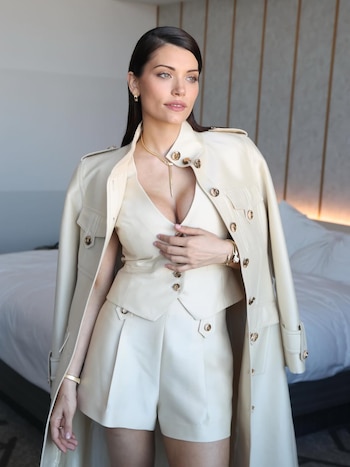 Eva De Dominici, de cabello oscuro, posa mirando a la derecha, vistiendo un conjunto crema de chaleco, shorts y abrigo, con joyas doradas