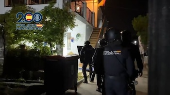 Varios agentes de policía con uniformes oscuros, cascos y escudos avanzan de noche hacia la entrada de una casa blanca de dos pisos