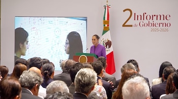 Claudia Sheinbaum habla desde un podio junto a la bandera de México, con una pantalla al fondo y público en primer plano.