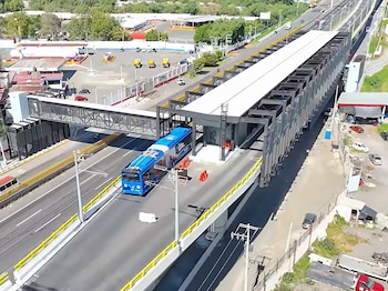 Vista aérea de una estación elevada de transporte con un vehículo azul articulado en la calzada y una autopista contigua