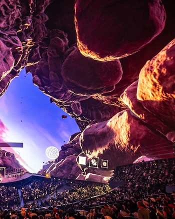 Vista interior de un concierto con una multitud sentada frente a un escenario con proyecciones en pantalla gigante de rocas y cielo azul