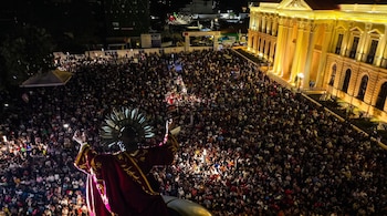 Gran multitud de personas congregadas de noche. Una estatua religiosa de espaldas, edificio con columnas iluminado, luces de teléfonos móviles y escenario.