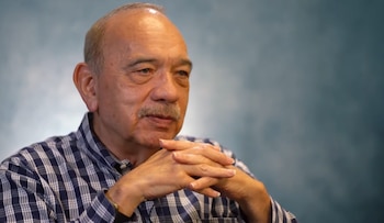 Un hombre calvo de edad avanzada con bigote, vistiendo una camisa de cuadros azul y blanco, con las manos entrelazadas, mira a la derecha