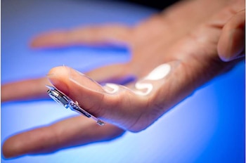 Mano con dedos extendidos sobre fondo azul, con un parche transparente en los dedos y palma, y un microchip metálico en la yema del dedo índice