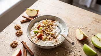Un tazón con yogur, trozos de manzana, nueces y avena sobre una mesa de madera rodeado de trozos de manzana y ramas de canela.