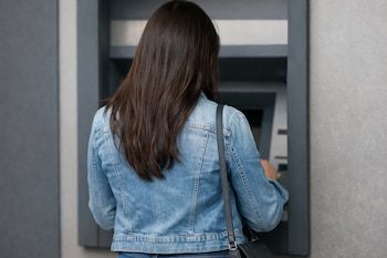 Mujer de espaldas con cabello castaño y chaqueta de mezclilla azul, usando un cajero automático gris. Su cuerpo cubre parcialmente el panel del cajero.