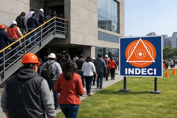 Grupo de personas evacua por escaleras exteriores de un edificio de concreto hacia una zona de césped, pasando un letrero azul con el logo de INDECI.
