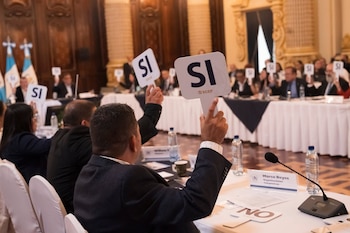 Varias personas en una mesa levantan carteles con la palabra SI. Al fondo, banderas de Guatemala. En primer plano, un micrófono y una placa con nombre