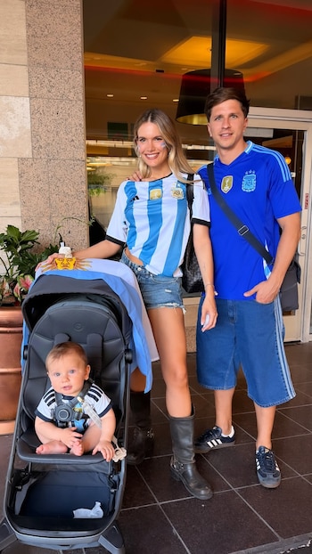 Eva Bargiela y Gianluca Simeone sonríen vestidos con camisetas de Argentina junto a su bebé en un cochecito, llevando una bandera albiceleste