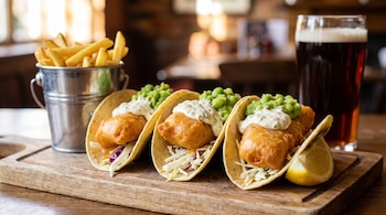 Tres tacos con pescado frito, salsa y guisantes, papas fritas en cubo, vaso de cerveza oscura y limón en una tabla de madera.