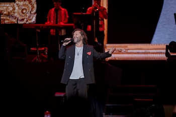 Diego Torres, con barba y cabello largo, canta en un micrófono en el escenario, vestido con saco oscuro y camiseta clara, con el brazo extendido y sonrisa. Músicos detrás