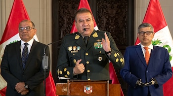 Un oficial en uniforme habla desde un podio flanqueado por dos hombres en traje y varias banderas de Perú.