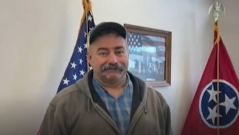 Hombre con bigote, gorra y chaqueta, de pie frente a las banderas de Estados Unidos y un estado, con un cuadro enmarcado detrás