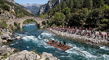Cuatro personas guían una balsa de troncos en un río rápido frente a una multitud reunida en la orilla y sobre un puente de piedra.