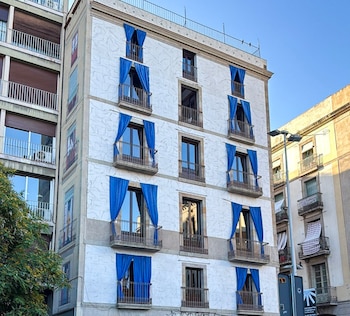 Fachada de un edificio blanco de siete pisos. Balcones con barandales de metal. Cortinas azules cubren las ventanas de varios balcones. Cielo azul