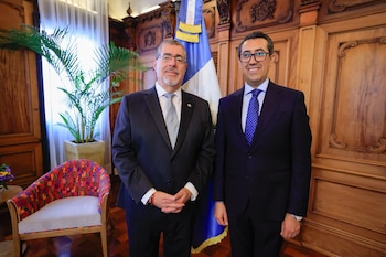 Dos hombres con traje formal se estrechan la mano ante la bandera de Guatemala en una habitación con paneles de madera