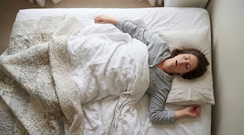 Vista aérea de una mujer durmiendo boca arriba en una cama con sábanas blancas y un edredón estampado, su boca está ligeramente abierta.