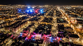 Un dron con luces encendidas vuela de noche sobre una ciudad con patrullas de policía estacionadas en una calle residencial.