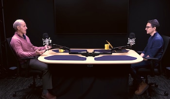 Dos hombres sentados frente a frente en una mesa con micrófonos profesionales, tazas amarillas y una computadora portátil en un estudio oscuro