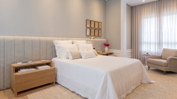Cama vestida de blanco con un amplio cabecero a cuadros, mesas de noche de madera, alfombra clara y un sillón junto a la ventana.