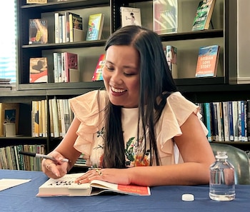 Mujer sonriente con camiseta floreada firma un libro sobre mesa con mantel azul. Al fondo, estanterías con libros y una botella de agua