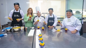 Cinco personas, una mujer de vestido verde claro y cuatro con uniformes de chef y delantales, decoran magdalenas con crema en una cocina