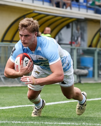 Un jugador de rugby masculino, con camiseta azul y blanca y pantalón corto blanco, corre con un balón ovalado en un campo de césped verde