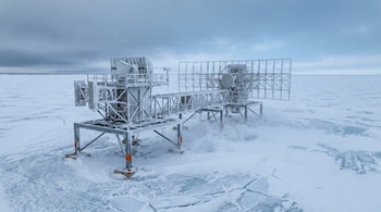 Estructura metálica con antenas y platos de radar sobre una superficie helada y agrietada bajo un cielo nublado.