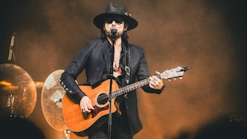 Beto Cuevas, el exvocalista de La Ley, en el escenario, usando un sombrero y gafas de sol, cantando en un micrófono y tocando una guitarra acústica