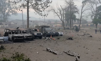 Vista de vehículos dañados, incluyendo varios SUV, en un terreno cubierto de escombros y ceniza con árboles y edificios al fondo bajo un cielo brumoso