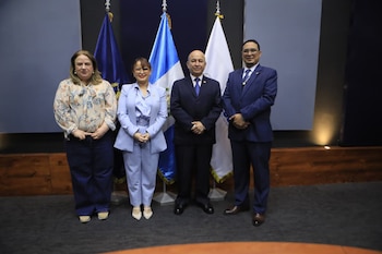Cuatro personas con atuendo formal, dos mujeres y dos hombres, posan frente a dos banderas de Guatemala y otras banderas lisas en un espacio interior iluminado