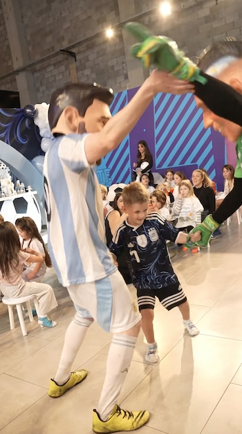 Un niño con camiseta de fútbol juega con dos personas disfrazadas. Una usa máscara de Messi y camiseta de Argentina, otra guantes de arquero. Hay niños sentados de fondo.
