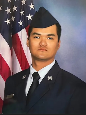 Retrato de un hombre con uniforme militar azul, gorra militar azul y corbata negra, frente a una bandera de Estados Unidos