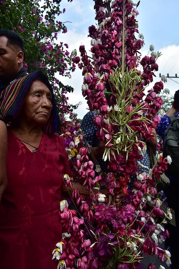 Primer plano de una mujer indígena mayor, de perfil, con tocado colorido y blusa roja, junto a un gran arreglo vertical de flores moradas y blancas