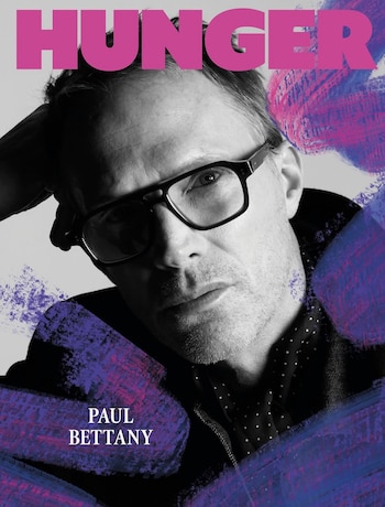 Retrato en blanco y negro de Paul Bettany con gafas, el texto HUNGER en la parte superior y trazos de pintura violeta en los bordes