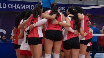 Siete jugadoras de voleibol con uniformes rojos y blancos se agrupan en una cancha de juego. Se ven números en sus espaldas y una red.