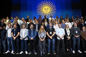 Un grupo diverso de personas de pie, en varias filas, frente a una pantalla azul con el Sol de Mayo amarillo de la bandera argentina