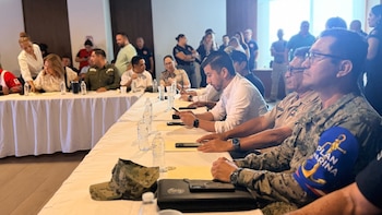 Personas vestidas con uniformes militares, de la Cruz Roja y ropa civil permanecen sentadas a lo largo de mesas blancas