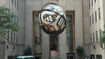 Escultura de Atlas sosteniendo un balón de fútbol gigante con texto ‘FIFA World Cup 2026’ frente a un edificio, con personas y vehículos en la calle
