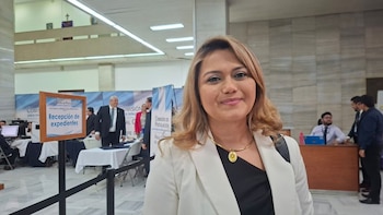 Mujer sonriente de cabello castaño claro con blazer blanco y collar dorado, en un entorno formal con banners de "Comisión de Postulación" de fondo