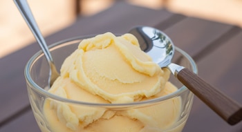 Nieve cremosa de color amarillo en un recipiente de vidrio transparente, con una cuchara de metal y otra con mango de madera sobre una mesa.