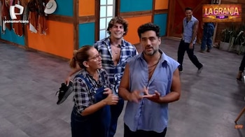 Tres personas, dos hombres y una mujer, interactúan en un set de televisión con paredes de madera. El hombre del centro gesticula, la mujer a su izquierda sostiene un zapato