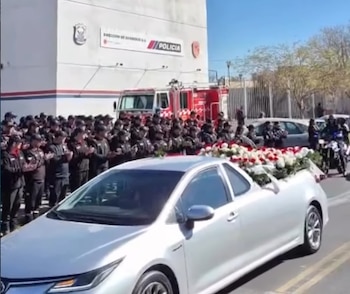 Agentes policiales y bomberos aplauden. Edificio con letreros de Policía y Bomberos. Coche fúnebre plateado con flores blancas y rojas. Camión de bomberos